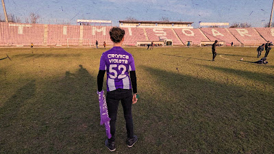 Un cronicar îmbrăcat în tricoul "Cronica Violeta" stă pe gazonul stadionului Dan Păltinișanu, privind spre tribunele aproape goale, în timp ce alți oameni se plimbă sau joacă fotbal.